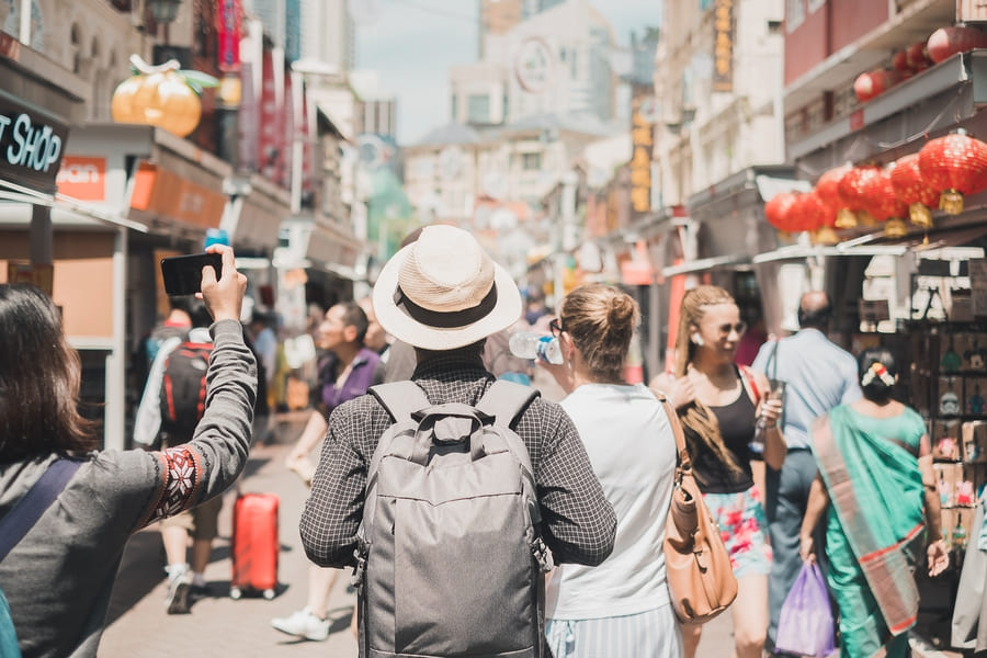 Turistas caminando de espaldas por una concurrida calle comercial de un Barrio Chino. Linternas rojas tradicionales adornan los locales bajo la luz de día en China